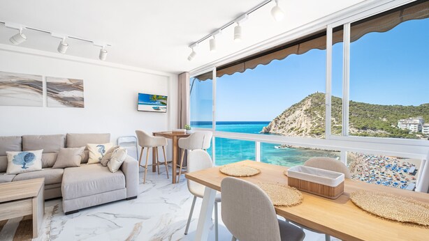 Een moderne woonkamer met een bank, eettafel en stoelen, grote ramen met uitzicht op zee en een berg.