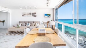 Een moderne woonkamer met een houten eettafel, een beige bank en grote ramen met uitzicht op de oceaan.