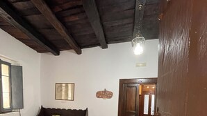 Una habitación con suelo de baldosas, puerta de madera, sofá con cojines, una lámpara de pared y una ventana con persianas.