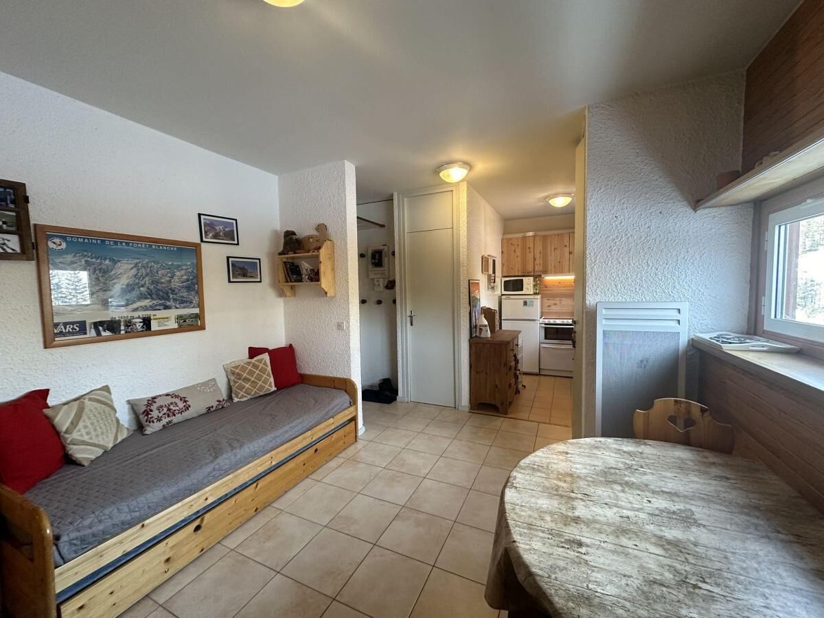 Quarto aconchegante com sofá de madeira, mesa de jantar e cozinha visível por uma porta aberta.