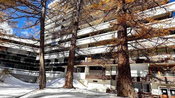 A multi-story building with a modern architectural design, surrounded by trees with autumn foliage and a snowy ground.