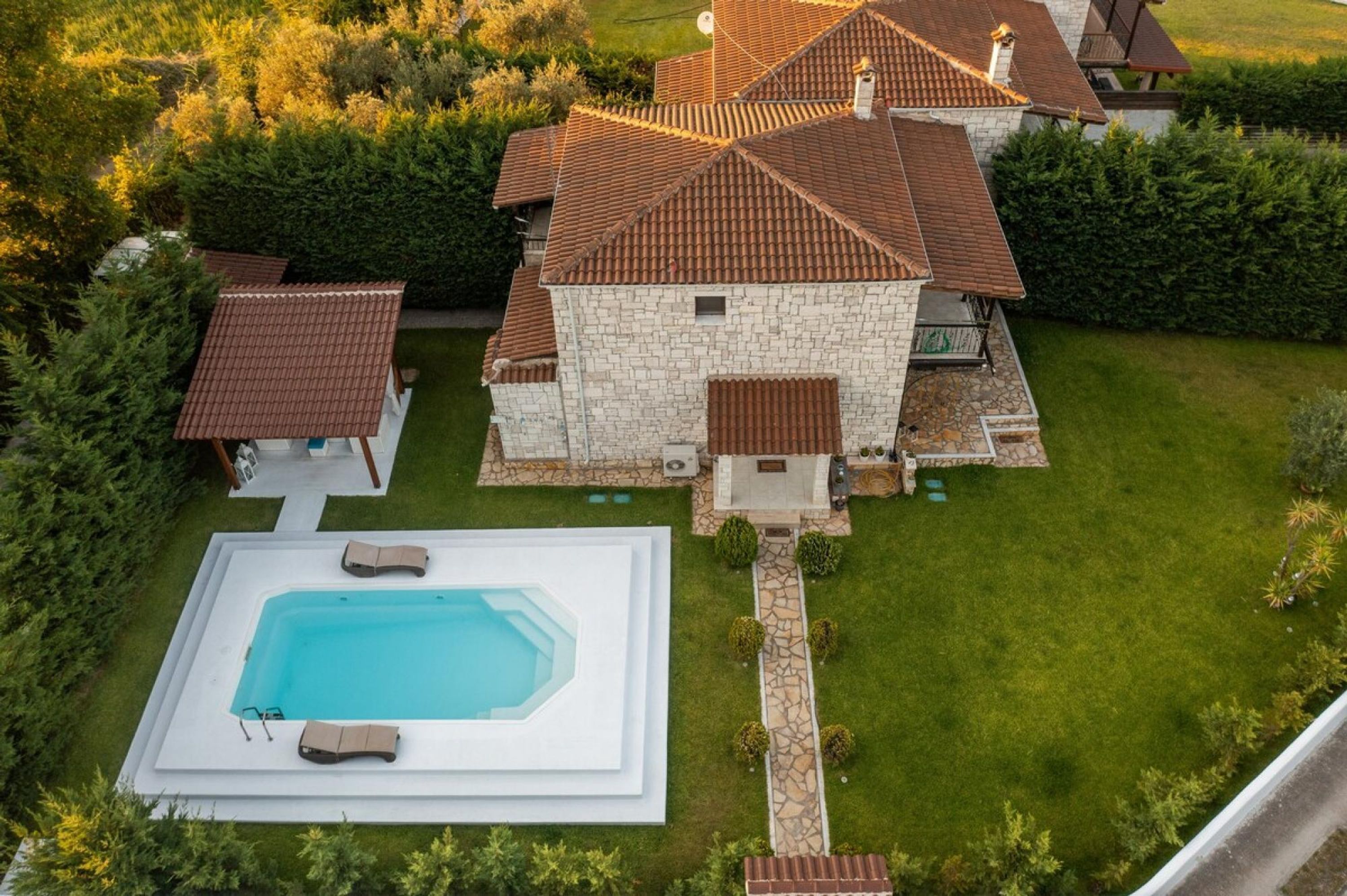 A large stone house with a swimming pool, surrounded by a well-maintained lawn and dense shrubbery.