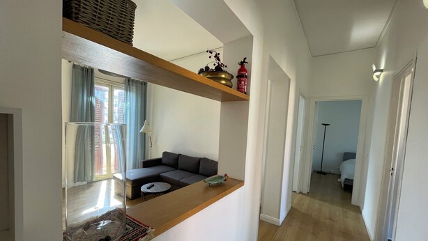 A modern living room with a sofa, a coffee table, and a small table with a decorative item. There is a window with curtains, a lamp, and a shelf with a basket and a fire extinguisher.