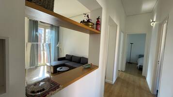 Un salon moderne avec un canapé, une table basse et une petite table sur laquelle est posé un objet décoratif. On y trouve une fenêtre avec des rideaux, une lampe et une étagère avec un panier et un extincteur.