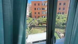 A view through a window with curtains, featuring a building with multiple balconies and air conditioning units, a fenced area with greenery, and a clear blue sky.
