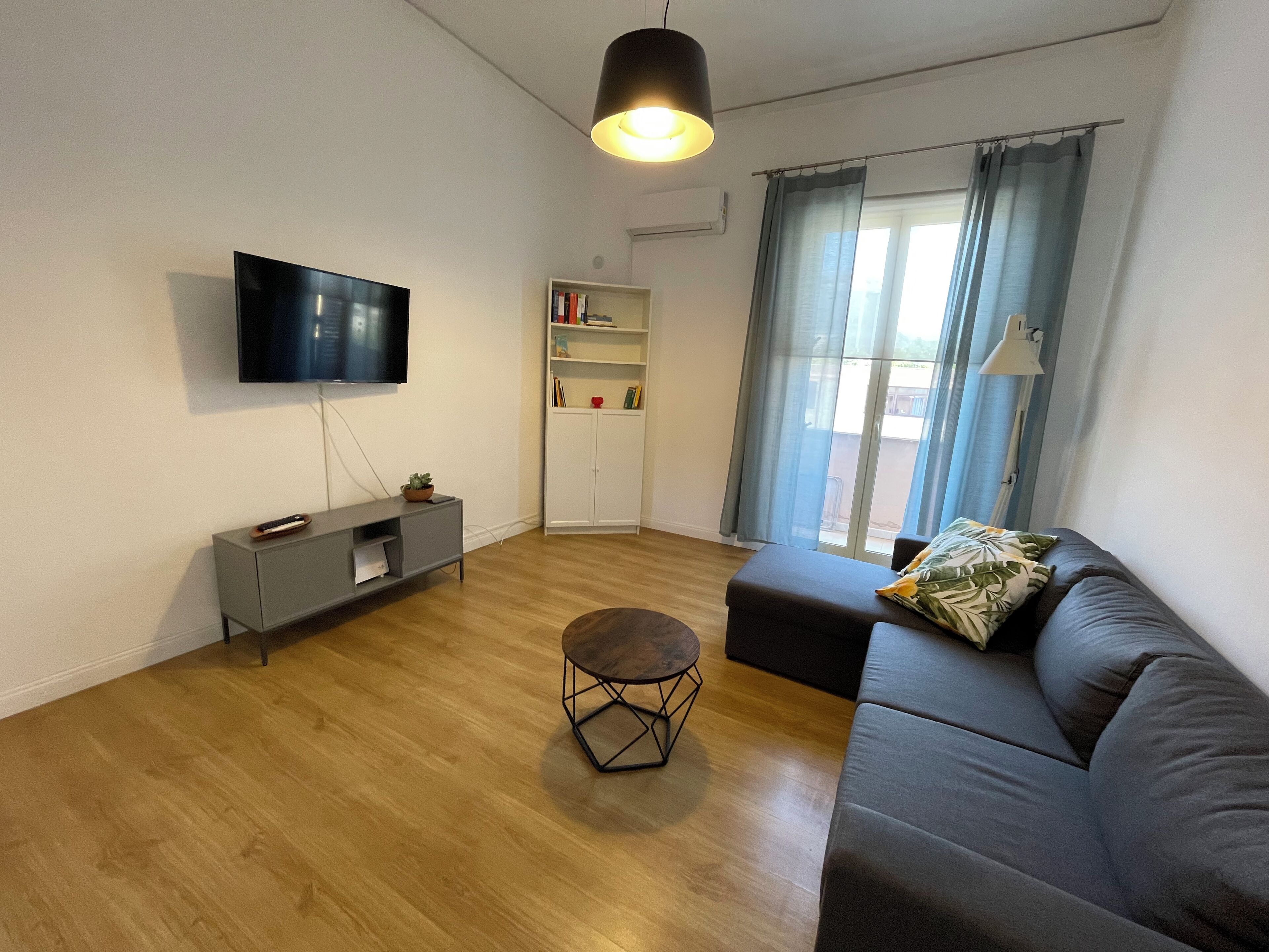 Un salon moderne avec un téléviseur à écran plat fixé au mur, un canapé gris, une petite table basse ronde et une bibliothèque.