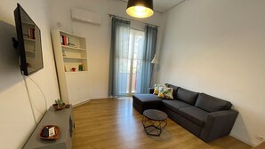 A modern living room with a grey sofa, a small round coffee table, a flat-screen TV mounted on the wall, a bookshelf, and a window with curtains.