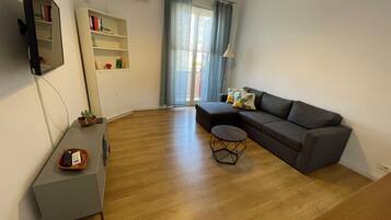 Un salon moderne avec un canapé gris, une petite table basse ronde, un téléviseur à écran plat fixé au mur, une bibliothèque et une fenêtre avec des rideaux.