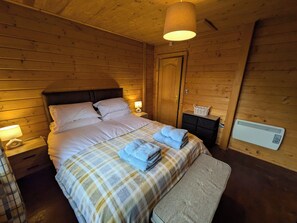 A wooden cabin bedroom with a bed, bedside lamps, a heater, and a bench. - Pet Friendly - Lodge at Riverview Holiday Park (Newcastleton)