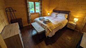 A wooden cabin bedroom with a bed, bedside tables, a bench, and a hanging light fixture. - Pet Friendly - Lodge at Riverview Holiday Park (Newcastleton)