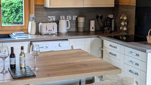 A compact kitchen with white cabinets, a wooden countertop, and various appliances including a toaster, kettle, and coffee maker. There is a window providing natural light and a view of greenery outside. - Pet Friendly - Lodge at Riverview Holiday Park (Newcastleton)