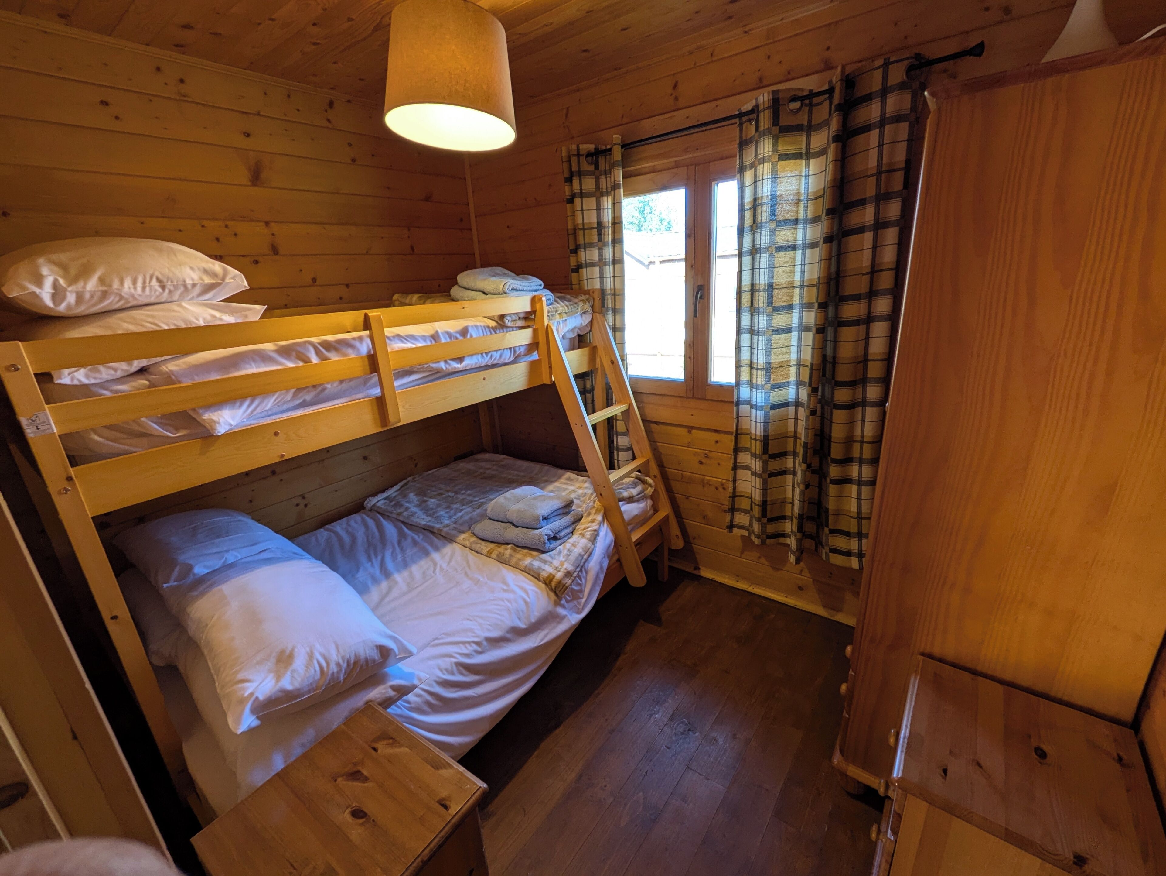 A wooden cabin room with bunk beds, a window with plaid curtains, and a wooden wardrobe.