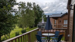 A wooden deck with a round table and chairs, overlooking a grassy area and trees. - Pet Friendly - Lodge at Riverview Holiday Park (Newcastleton)