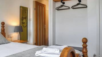 Habitación de hotel con una cama, cabecera de madera, mesita de noche con lámpara, espejo y una puerta que da a otra habitación.