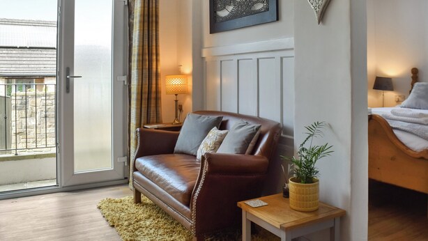 Un salon chaleureux avec un canapé en cuir marron, une petite table d’appoint en bois, une plante en pot et une porte-fenêtre coulissante donnant sur un balcon.