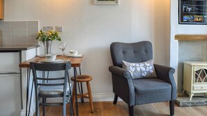 Un coin repas avec une table dressée pour deux personnes, un fauteuil à revêtement gris agrémenté d’un coussin à motifs floraux, et une œuvre d’art encadrée accrochée au mur.
