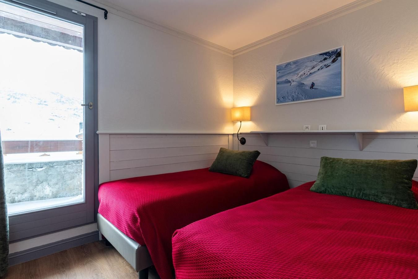 A room with two beds, each with a red bedspread and a green pillow. There is a window on the left side, and a wall-mounted picture above the beds.