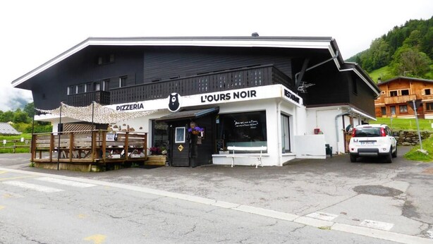 Una pizzería llamada 'L'Ours Noir' con fachada blanco y negro, un área de asientos al aire libre de madera y un coche blanco estacionado en la calle.