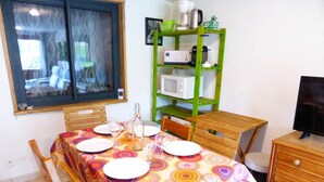 Un comedor con una mesa preparada para cuatro, un estante verde con electrodomésticos de cocina y un televisor sobre un mueble de madera.