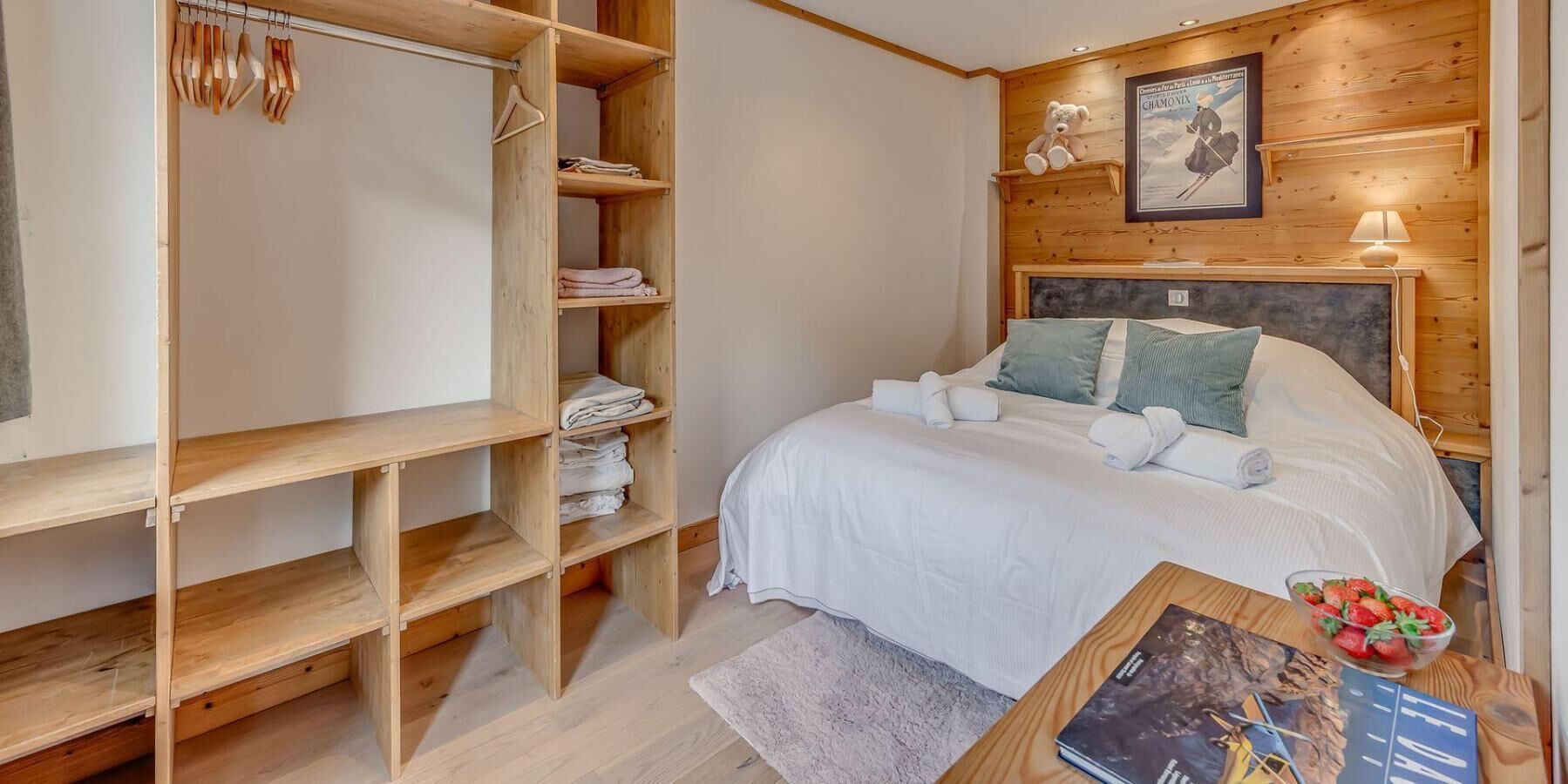 A bedroom with a wooden wardrobe, a bed with white bedding, a small table with a book and a bowl of fruit, and a framed picture on the wall.