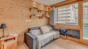 A cozy room with a wooden wall, a sofa, a small table with a lamp, and a window with a view of buildings. - 1st floor, tv, ski locker, 50m², Tignes (Tignes)