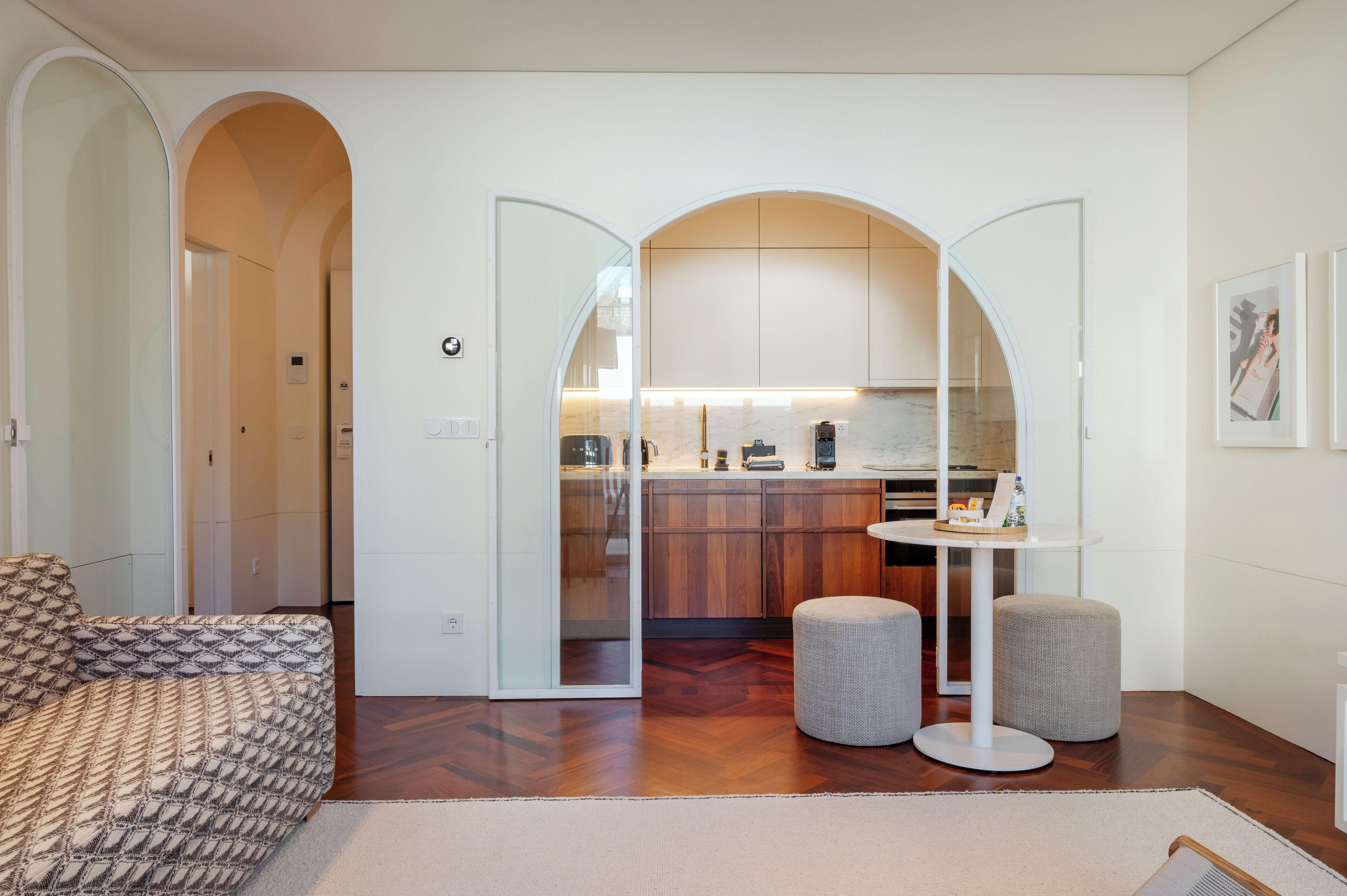Un salon moderne avec un canapé à motifs, une table ronde accompagnée de deux poufs, et une cuisine visible à travers une arche.