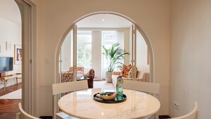 Un comedor con una mesa de mármol, sillas blancas y vistas a un salón con sofá, planta y televisor, a través de un arco.