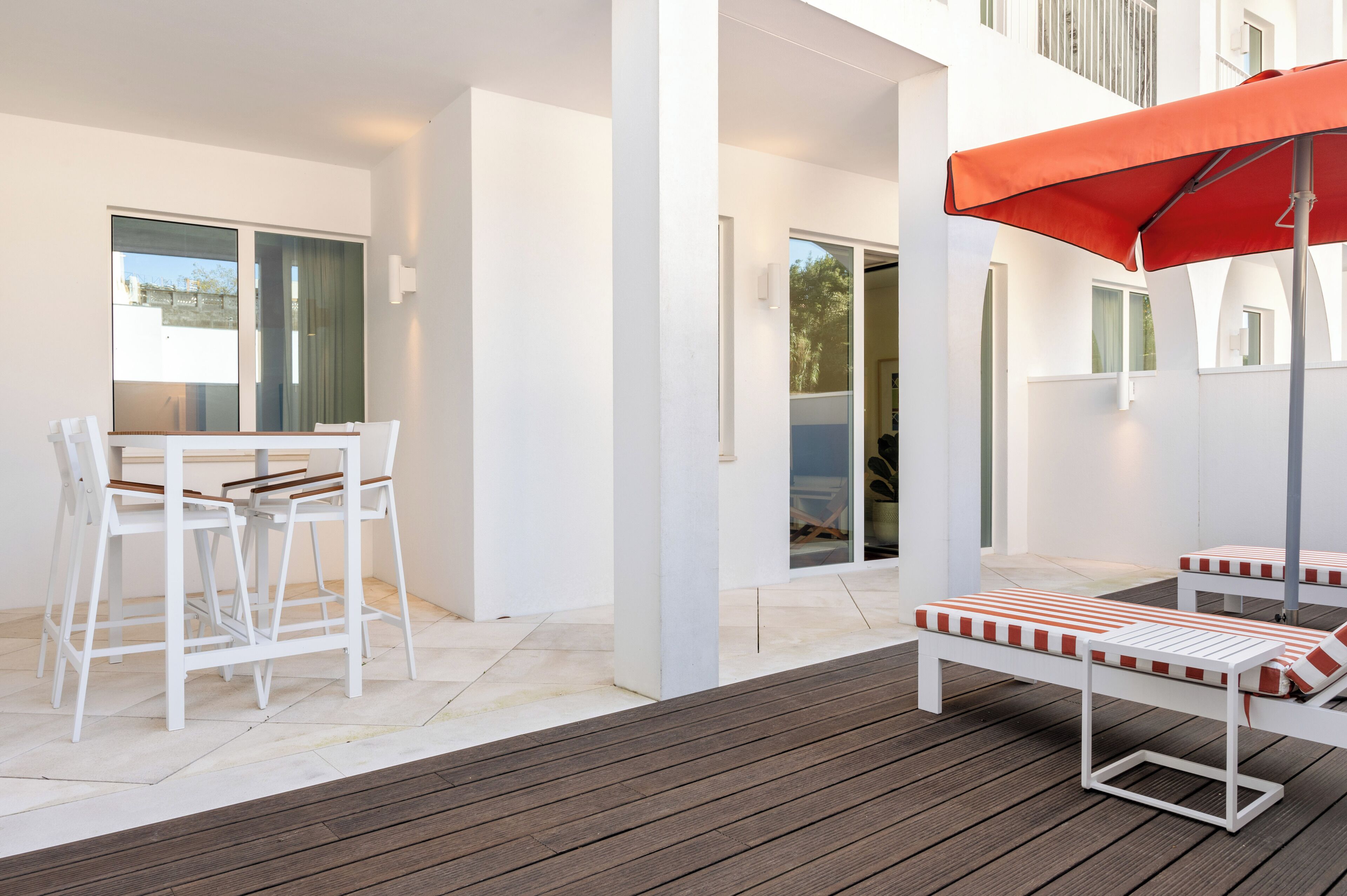 Un espace extérieur moderne, aménagé avec des chaises blanches et une table, une terrasse en bois et un parasol rouge.