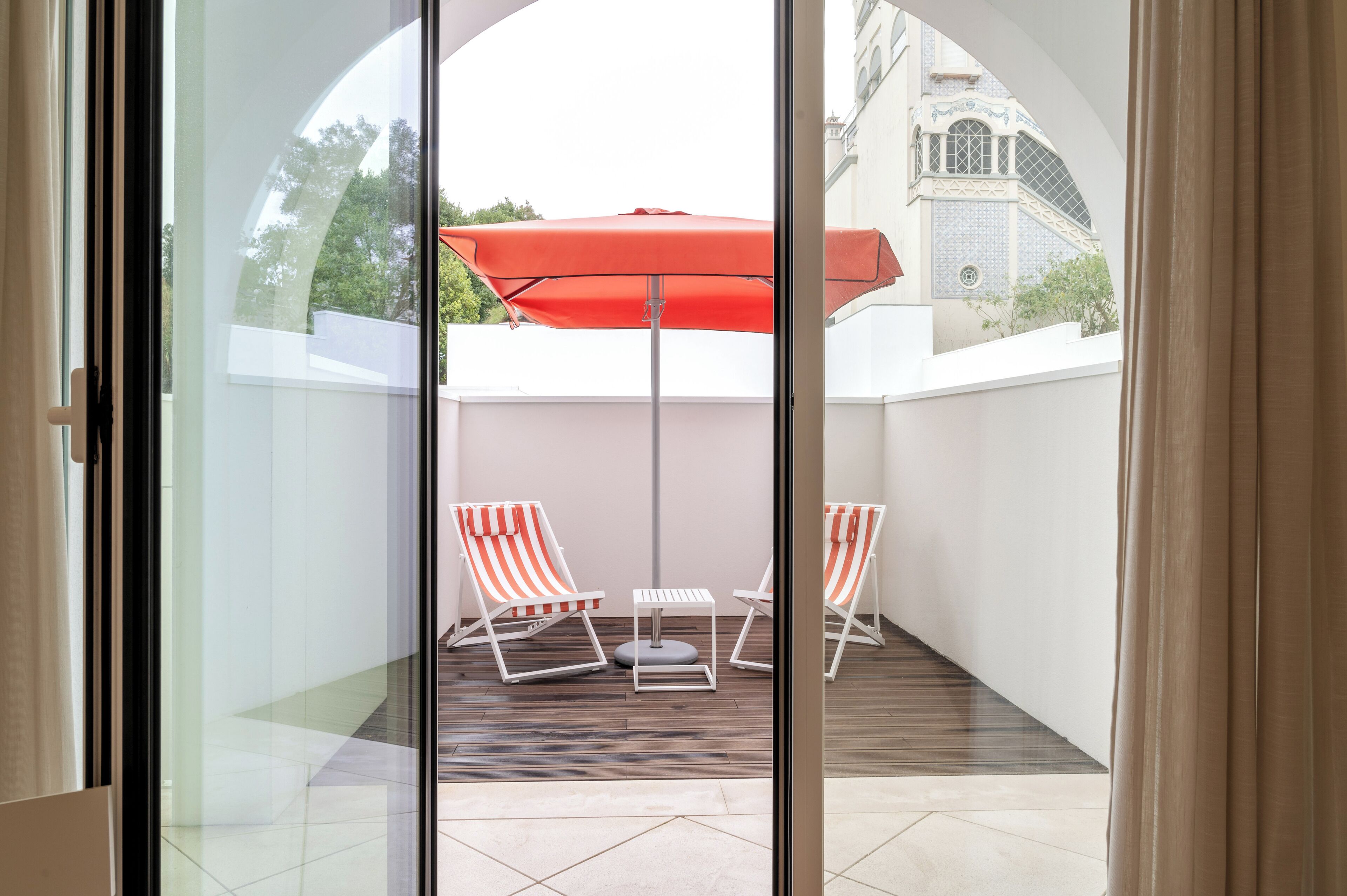 Uma porta de vidro dá para uma varanda com duas cadeiras listradas, uma mesa pequena e um guarda-sol vermelho.