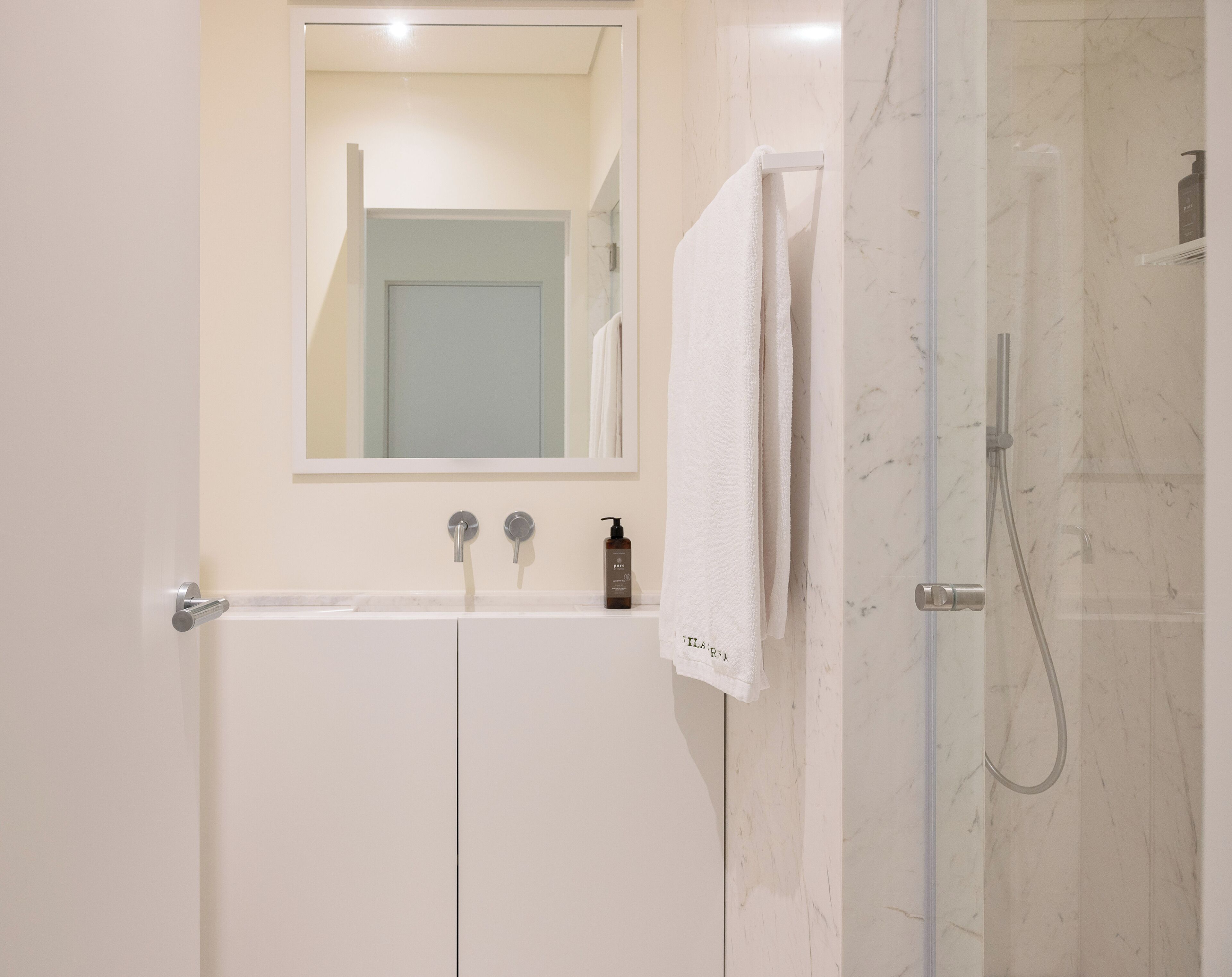 Une salle de bain avec un lavabo blanc, deux robinets, un miroir, un porte-serviettes et une douche à parois de verre.