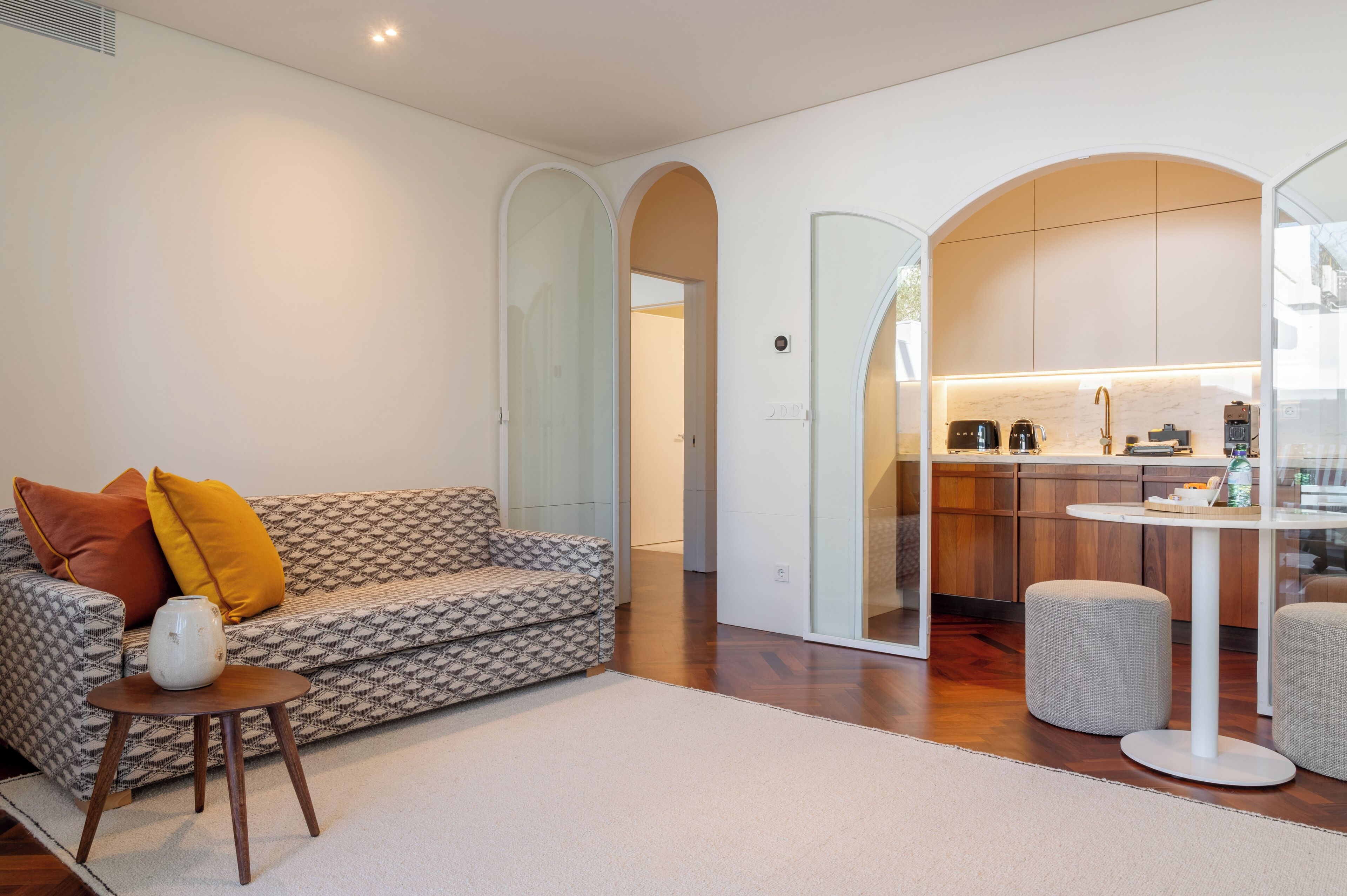 A modern living room with a patterned sofa, a small round table, and a kitchen area with a white countertop and wooden cabinets.
