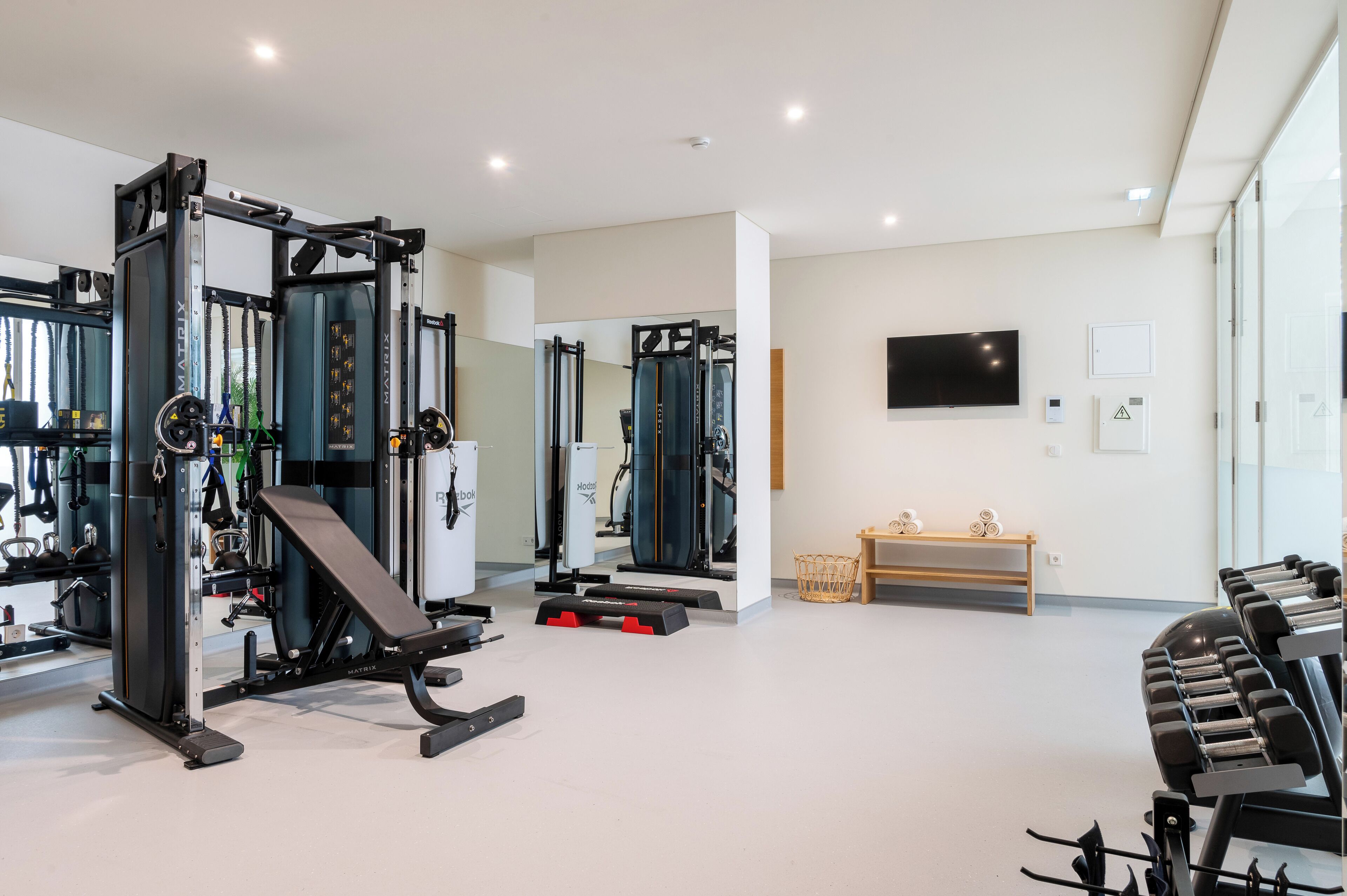 A modern gym with various exercise equipment, a television on the wall, and a bench with dumbbells.