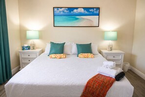 A hotel room with a large bed, two nightstands, a wall-mounted painting of a beach, and a teal curtain.