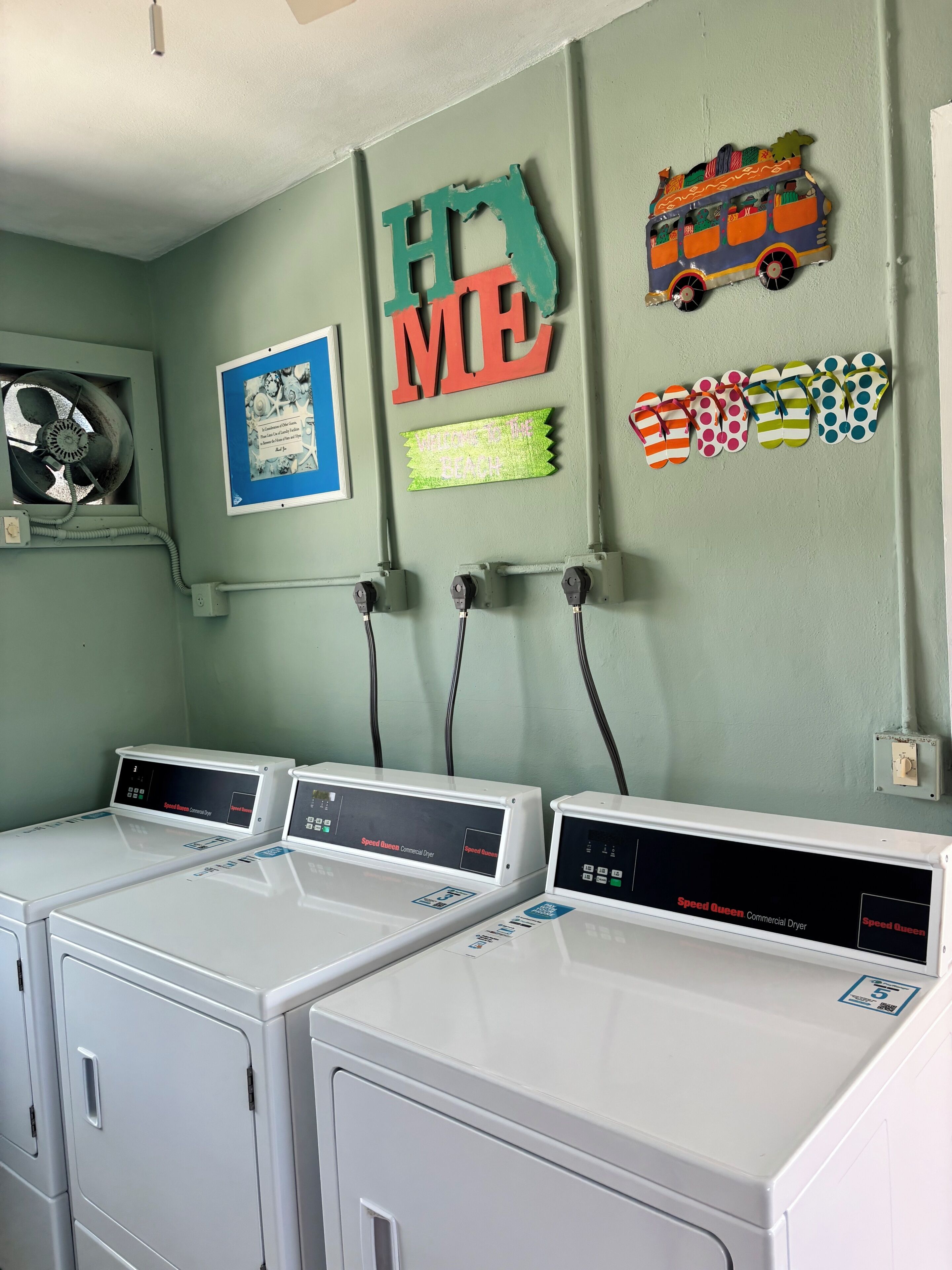 A laundry area with three Speed Queen dryers, a wall-mounted fan, and decorative items including a bus and a sign that reads 'WELCOME TO THE BEACH.'