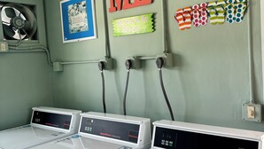 Zona de lavandería con tres secadoras Speed Queen, un ventilador de pared y objetos decorativos, incluyendo un autobús y un letrero que dice 'BIENVENIDOS A LA PLAYA'.