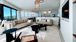 Ein modernes Wohnzimmer mit einer großen Couch, einem Couchtisch und einem Essbereich. Große Fenster bieten einen Ausblick ins Freie.