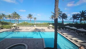A pool area with lounge chairs and umbrellas, surrounded by palm trees and a view of the ocean.