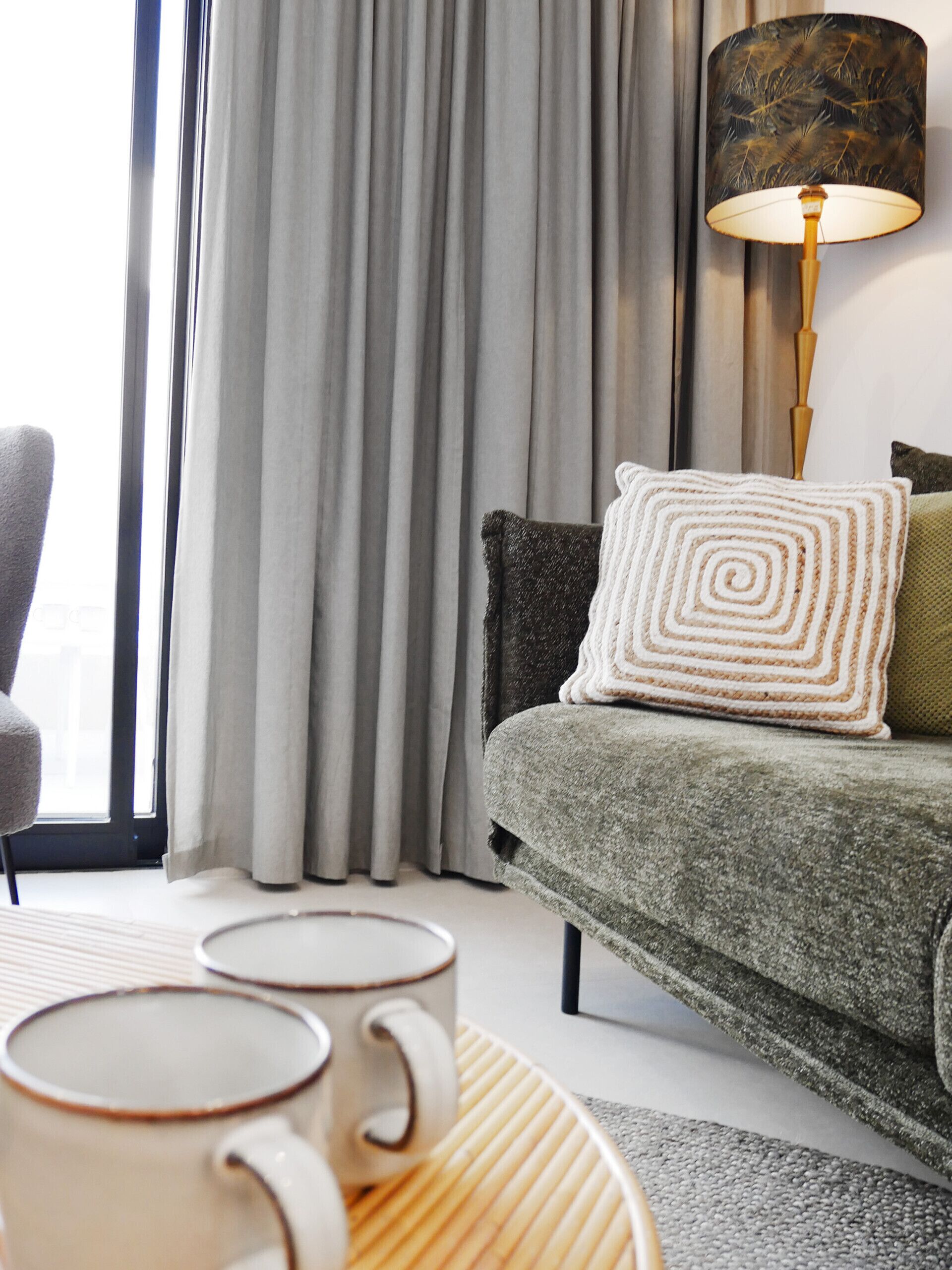 A modern living room with a grey sofa, a patterned throw pillow, a lamp, and two mugs on a woven tray.