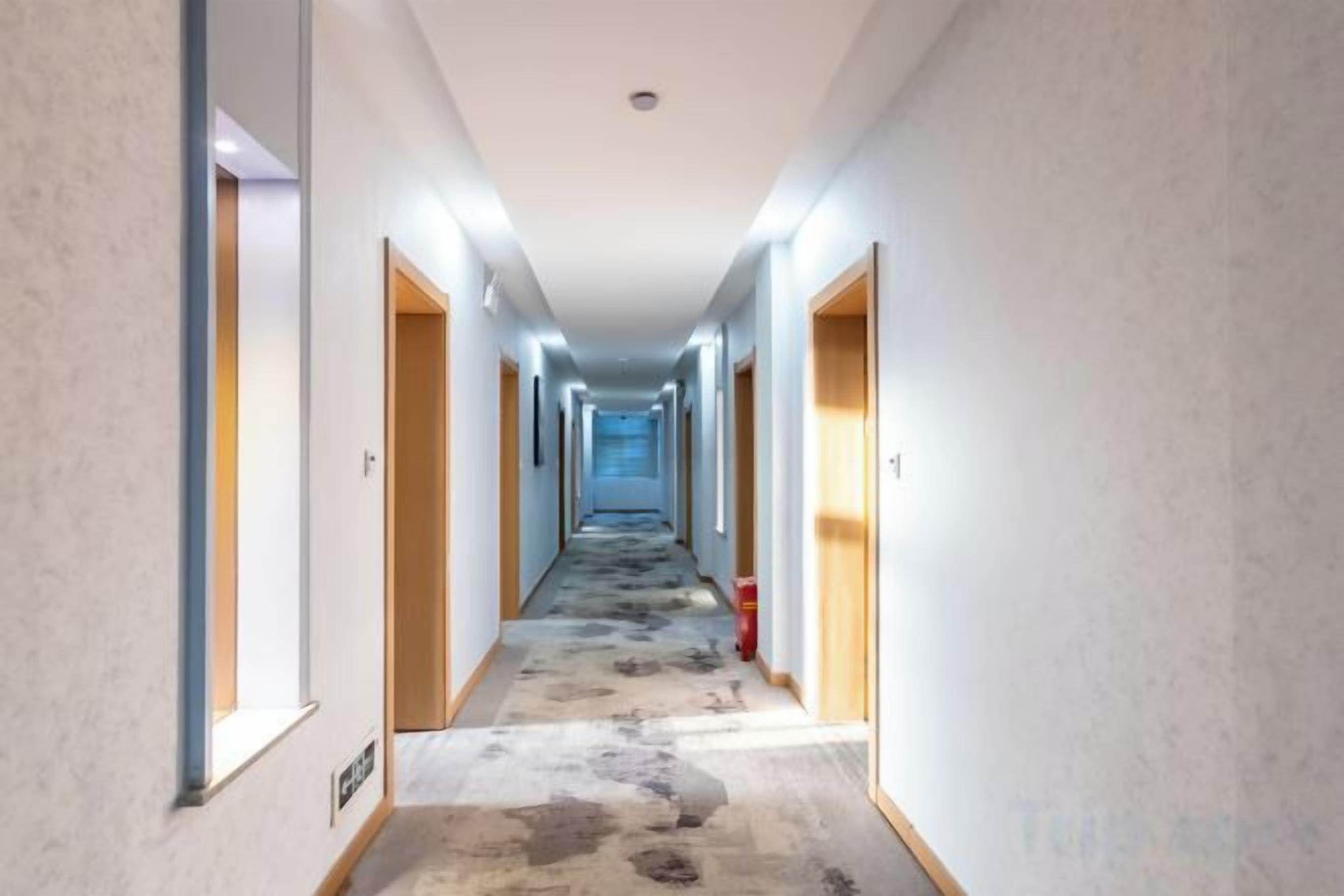 En hotellkorridor med dörrar på båda sidor, ett mönstrat golv och en brandsläckare på höger sida.