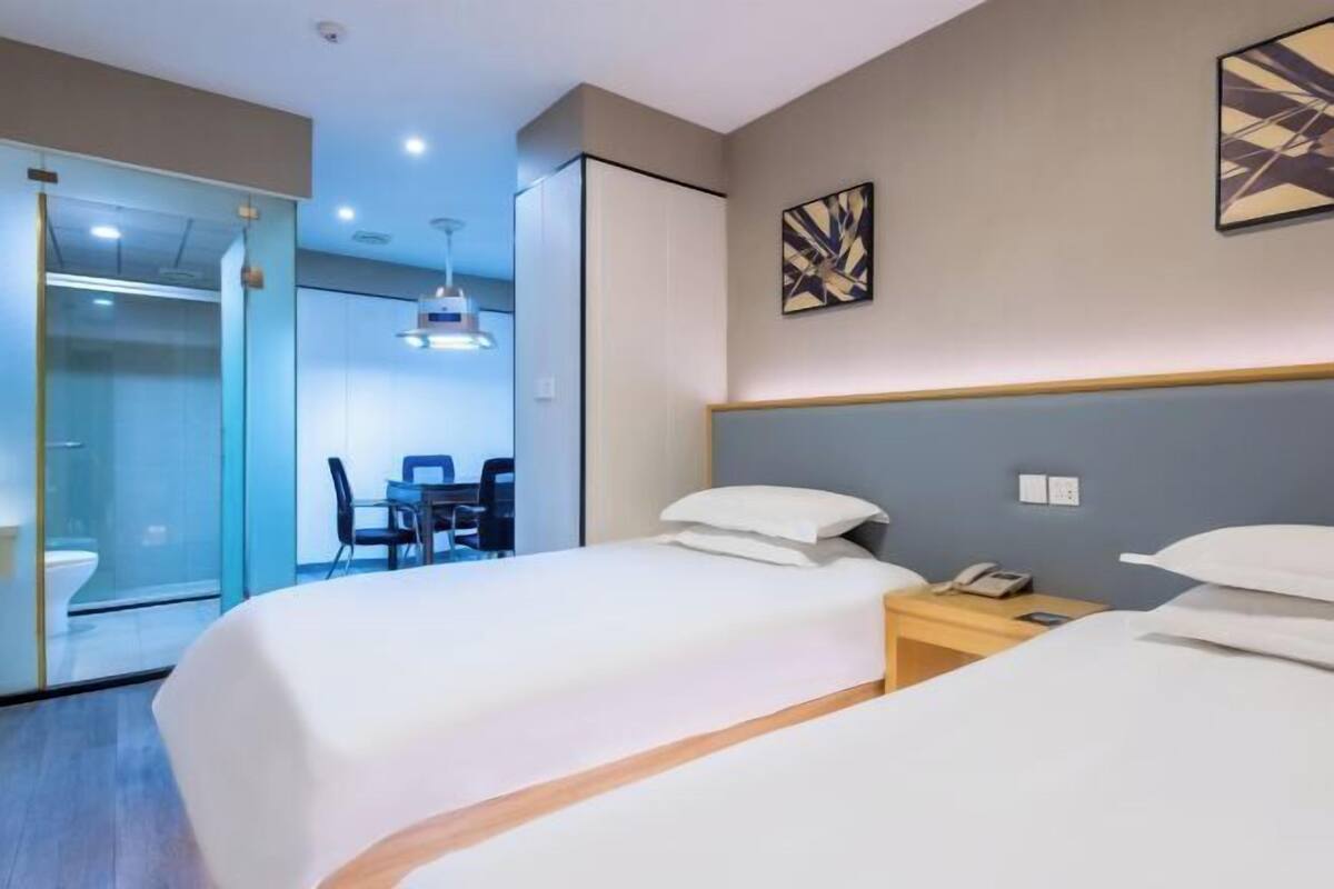A hotel room with two beds, a desk, and a glass partition separating the bathroom.