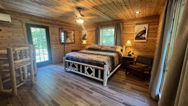 Ein Schlafzimmer einer Holzhütte mit einem Bett, zwei Fenstern, einem Deckenventilator und einem Holzstuhl.