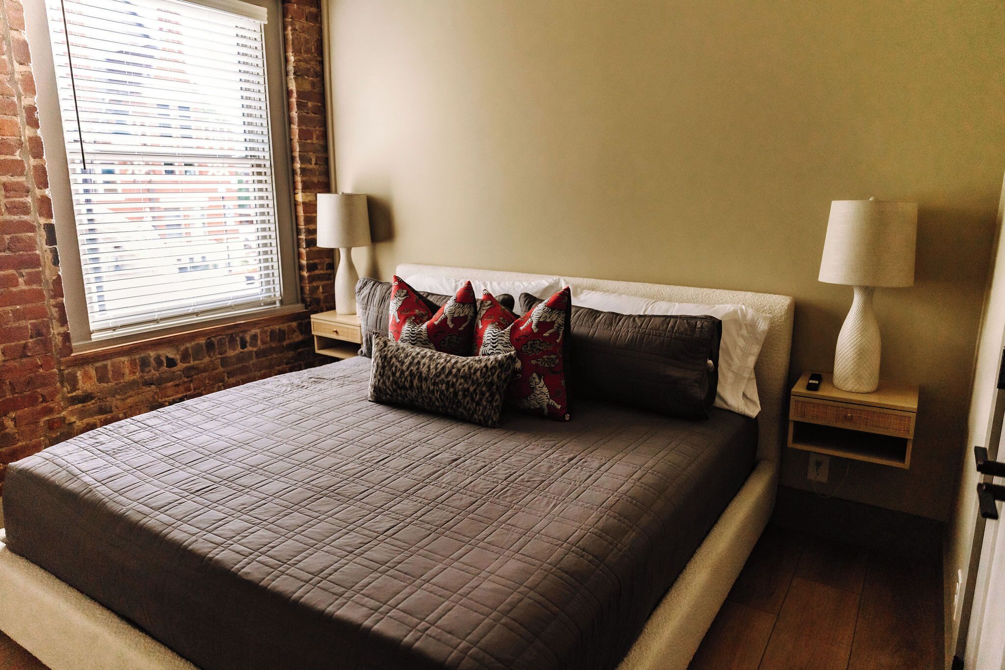 Een slaapkamer met een bakstenen muur, een raam met zonwering, een bed met een gewatteerde sprei, twee bedlampen en een nachtkastje met een lamp.