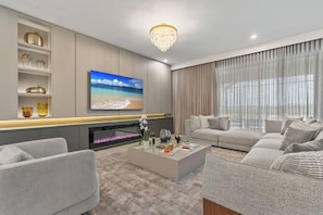 Welcome to a cozy living room featuring plush gray sofas perfect for relaxation. A modern chandelier and sleek electric fireplace add warmth, while large windows with elegant curtains provide a lovely view of the outside.