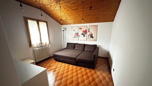 A room with a wooden ceiling, a grey sofa, a radiator, and a window with curtains.