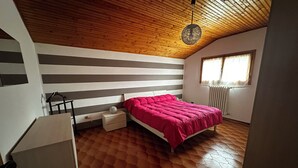 A bedroom with a wooden ceiling, a bed with a pink blanket, a nightstand, a dresser, and a window with curtains.