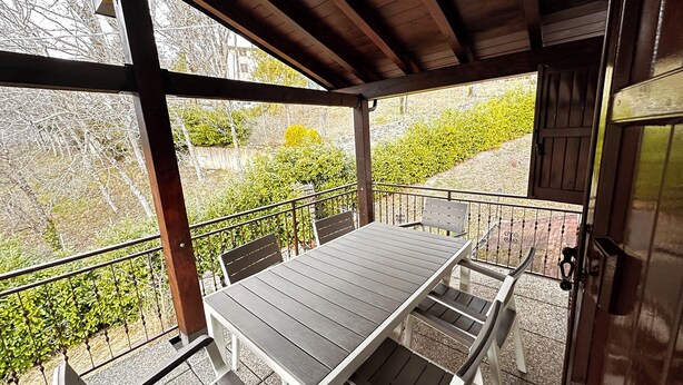 A covered outdoor seating area with a table and chairs, a railing, and a view of greenery and buildings.