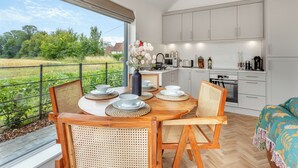 A modern kitchen with white cabinets, a dining area with a round table and chairs, and a view of a green field through large windows. - UK47693 - Piglet Cottage (Wimbish)