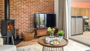 A cozy living room with a brick wall, a fireplace, a flat-screen TV, a dining table with a plate of food, and a large window. - UK47693 - Piglet Cottage (Wimbish)