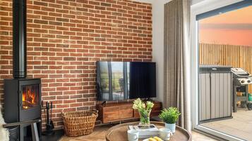 A cozy living room with a brick wall, a fireplace, a flat-screen TV, a dining table with a plate of food, and a large window.