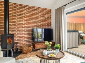 A cozy living room with a brick wall, a fireplace, a flat-screen TV, a dining table with a plate of food, and a large window.